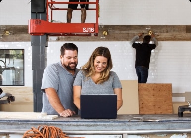 Owners of a construction business look over reports and invoices while their crew works in the background.