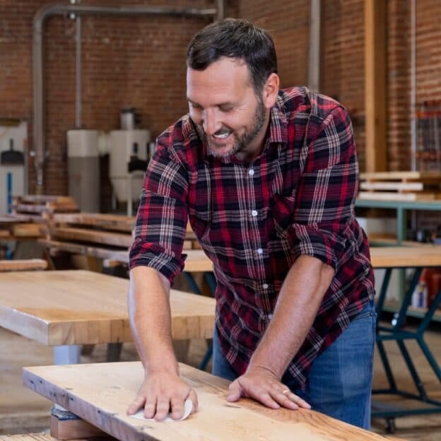 A photo of a person in a woodworking shop leaning over a plank of wood as they sand it. The person is smiling as they sand the wood. Behind them you can pieces of furniture being built and an exposed brick wall.